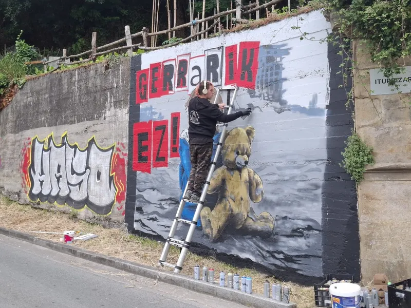 Artista sobre una escalera pintando el mural Gerra rik ez